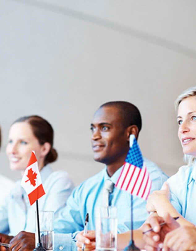 Two Caucasian women and a black man seated behind a Canadian and a U.S. flag at a meeting table, smiling.