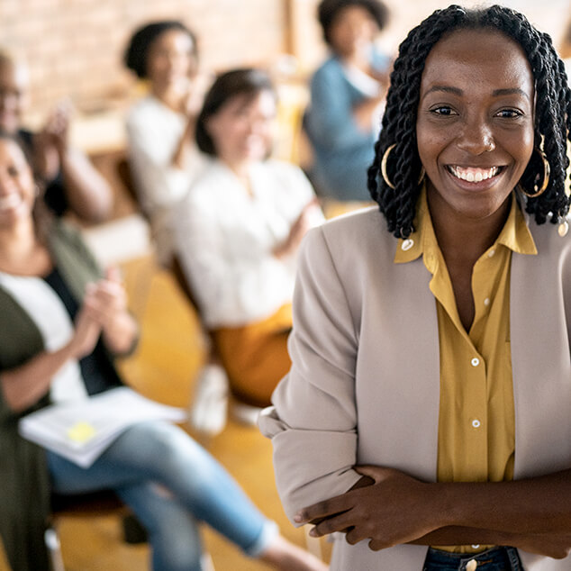 A smiling young professional woman in the foreground with a group of young professionals seated behind her. 