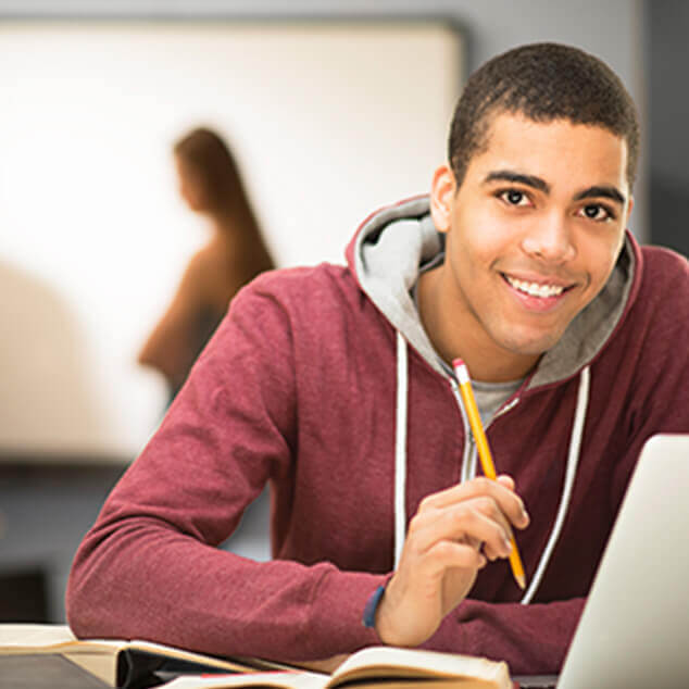 A smiling college-age male student using a laptop and studying with a book and pencil in hand. 
