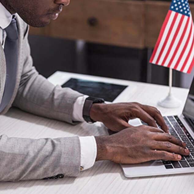 A black businessman typing on a laptop with an American flag on a desk. 