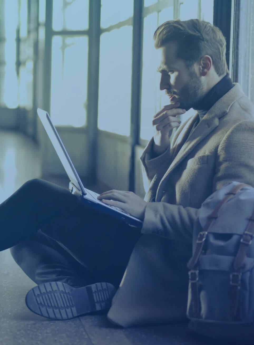 A Caucasian man seated on the floor with his laptop, looking pensively at the screen.