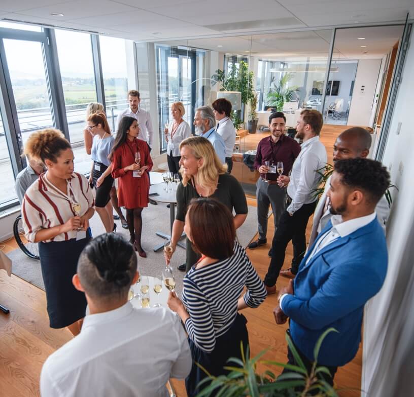 A diverse group of professionals standing up, talking, and networking in a modern meeting venue. 