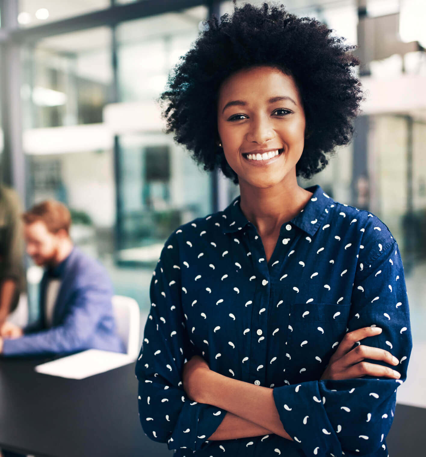 A professional woman smiling with crossed arms in the foreground and two men working at a desk in the background in a modern office. 