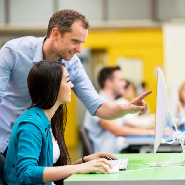 A male teacher leaning over a female adult student who is working on a desktop computer. 