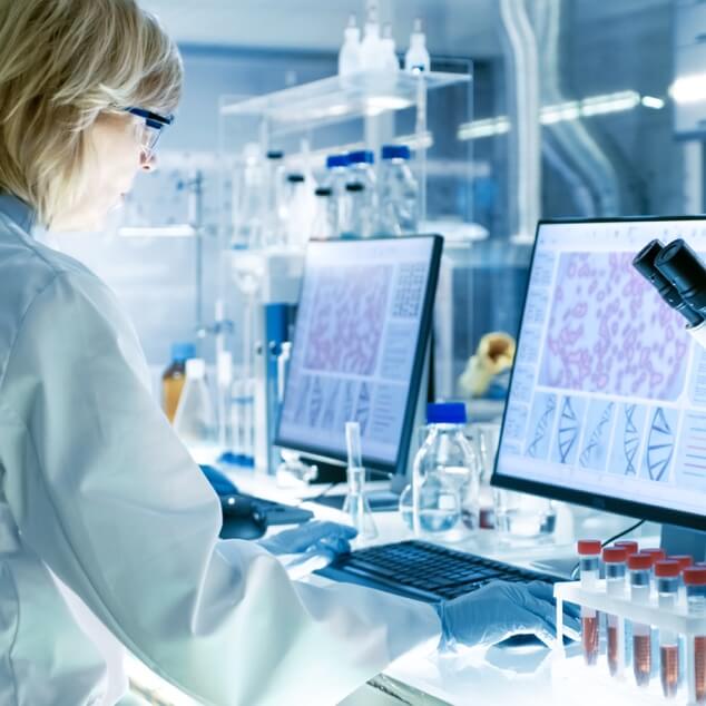 A researcher in a white coat looking at microscopic scan images on a computer monitor.