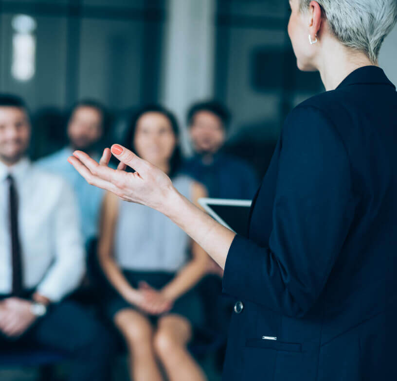 A Caucasian woman speaking in front of a group of seated professionals. 