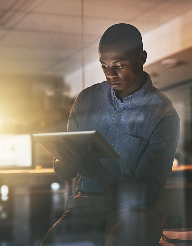 A young man sitting looking at a table computer with sunlight streaming into a dark room. 