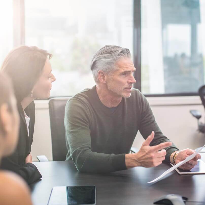 An older Caucasian man seated at a table with a tablet computer, speaking to a group at the table. 