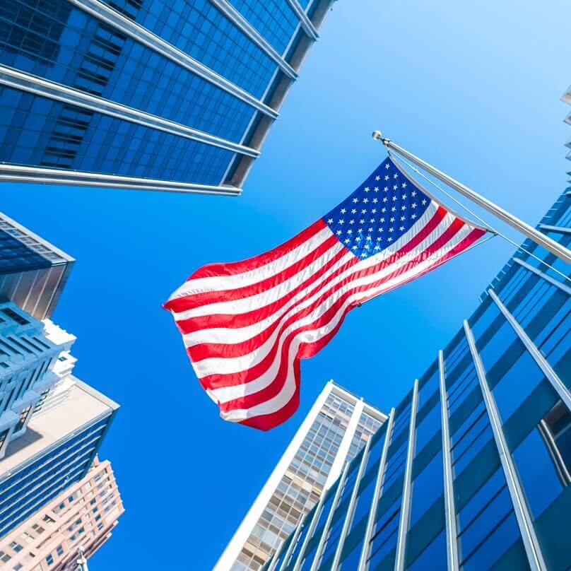 Upward view of an American flag flying against a bright blue sky in front of modern buildings.