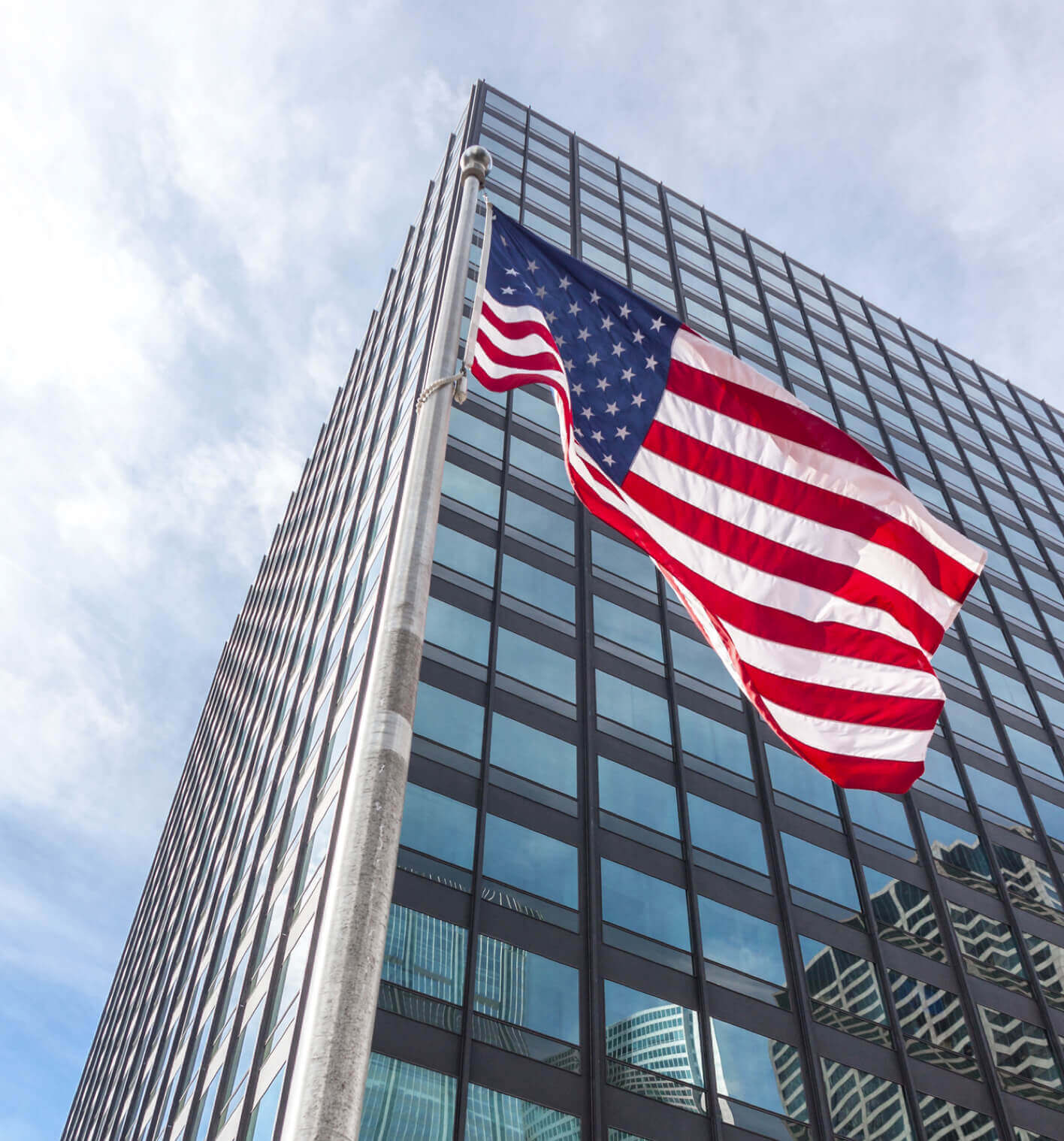 An upward view of an american flag flying in front of a modern office building and a bright sky.